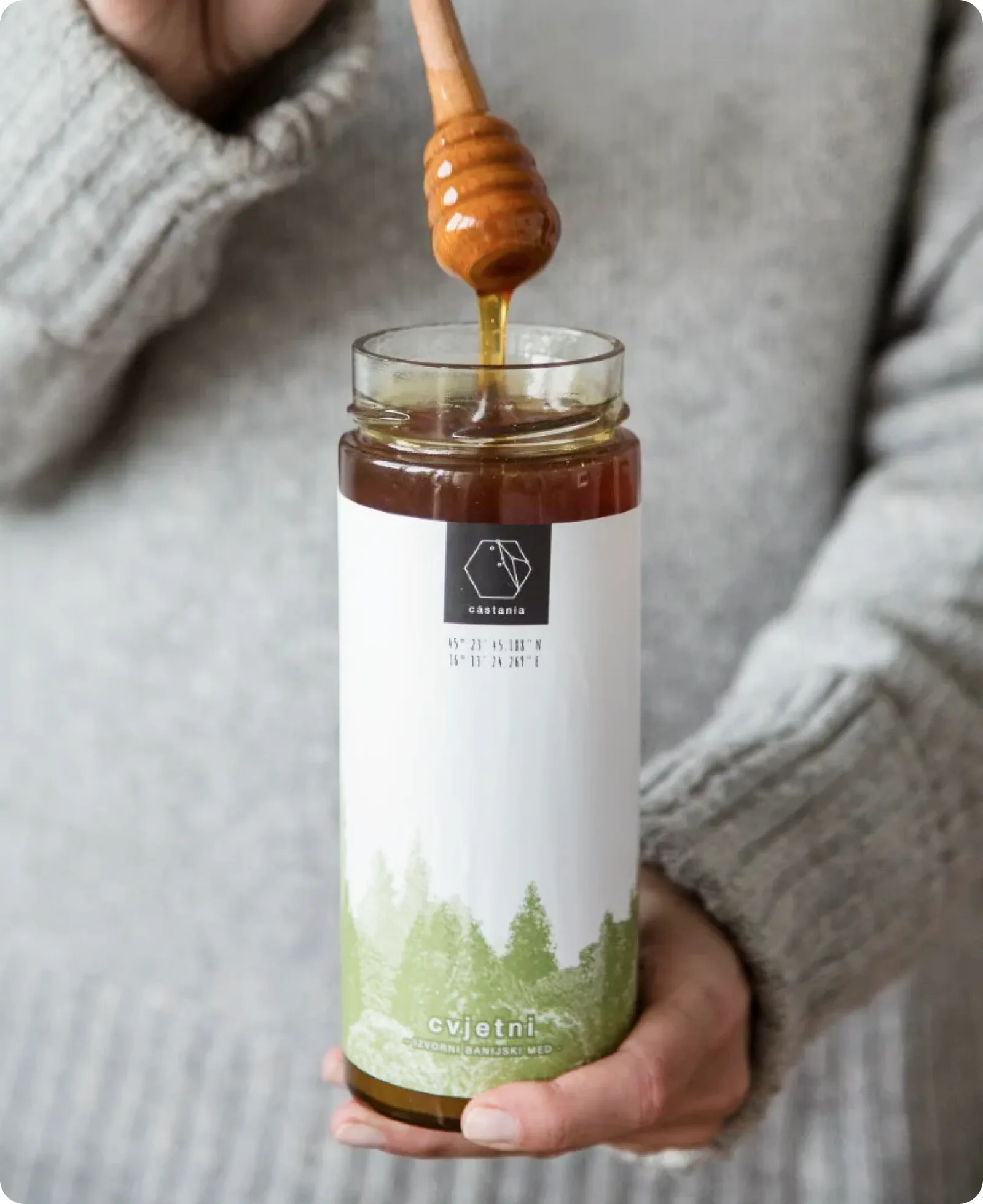 A hand holding a Castania honey jar while honey drips from a wooden dipper.