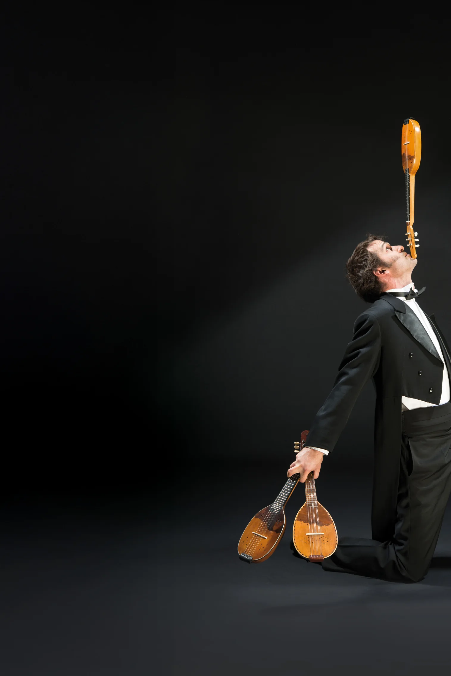 A tamburitza musician in white tie kneeling, balancing a bisernica on his chin and holding two more instruments.