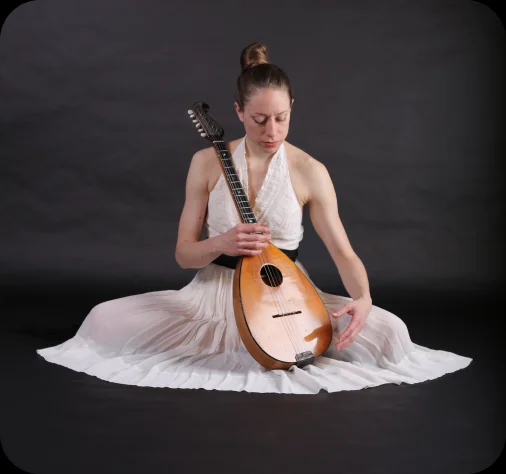 A dancer in a white dress seated on the floor, cradling a large tamburitza.